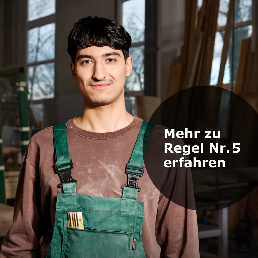 Junger Mann in grüner Arbeitslatzhose mit einem Klappmaß in der Tasche steht in einer Werkstatt