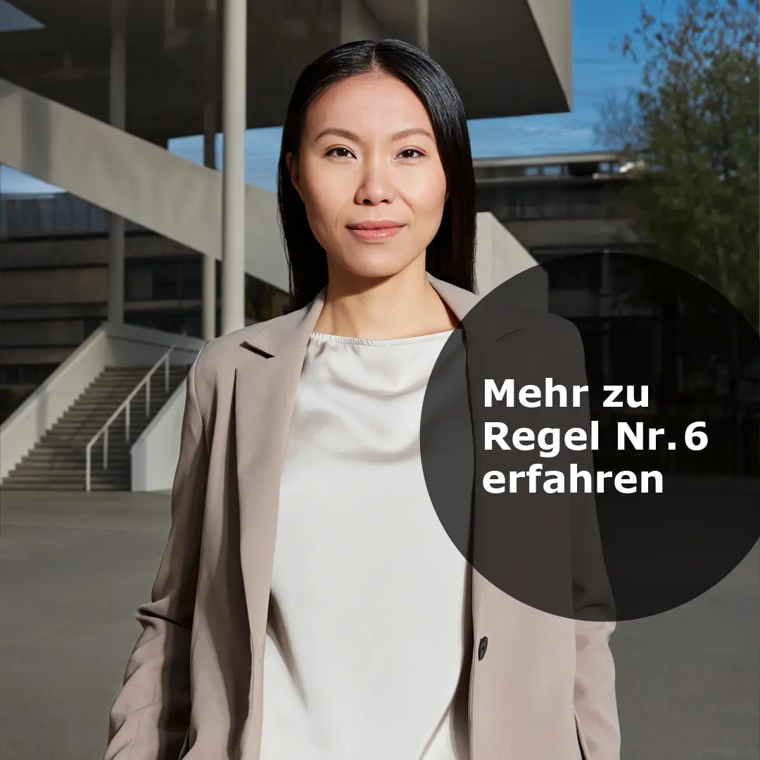 Frau mit langem schwarzen Haar in beigem Blazer und weißer Bluse vor einer modernen Treppe im Hintergrund.