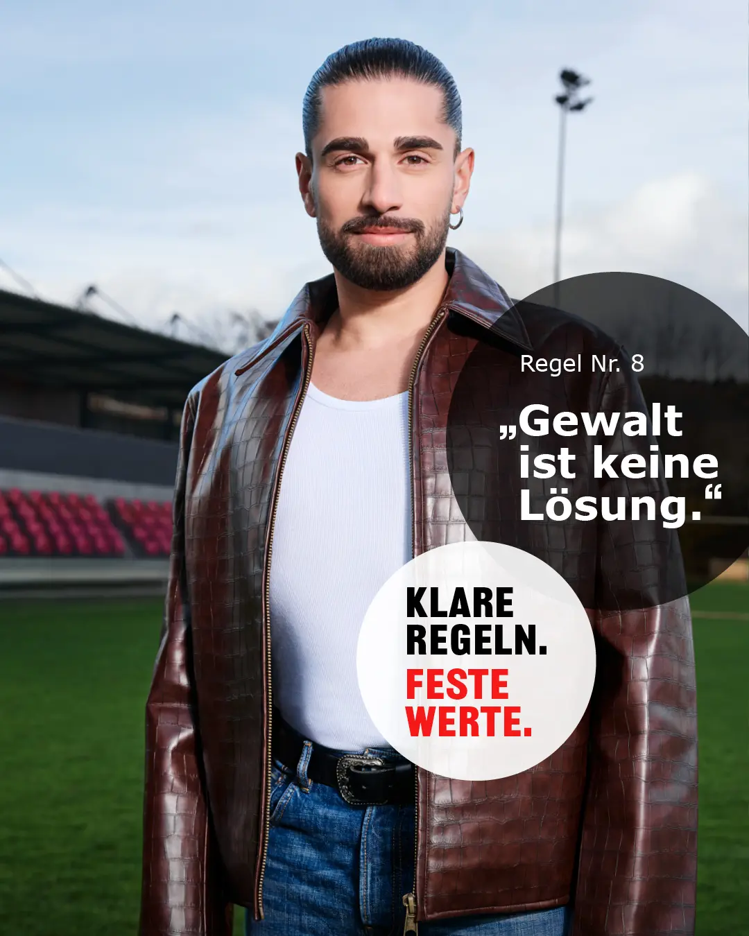 Mann mit Lederjacke und zusammengebundenen Haaren steht auf einem Fußballfeld mit Text