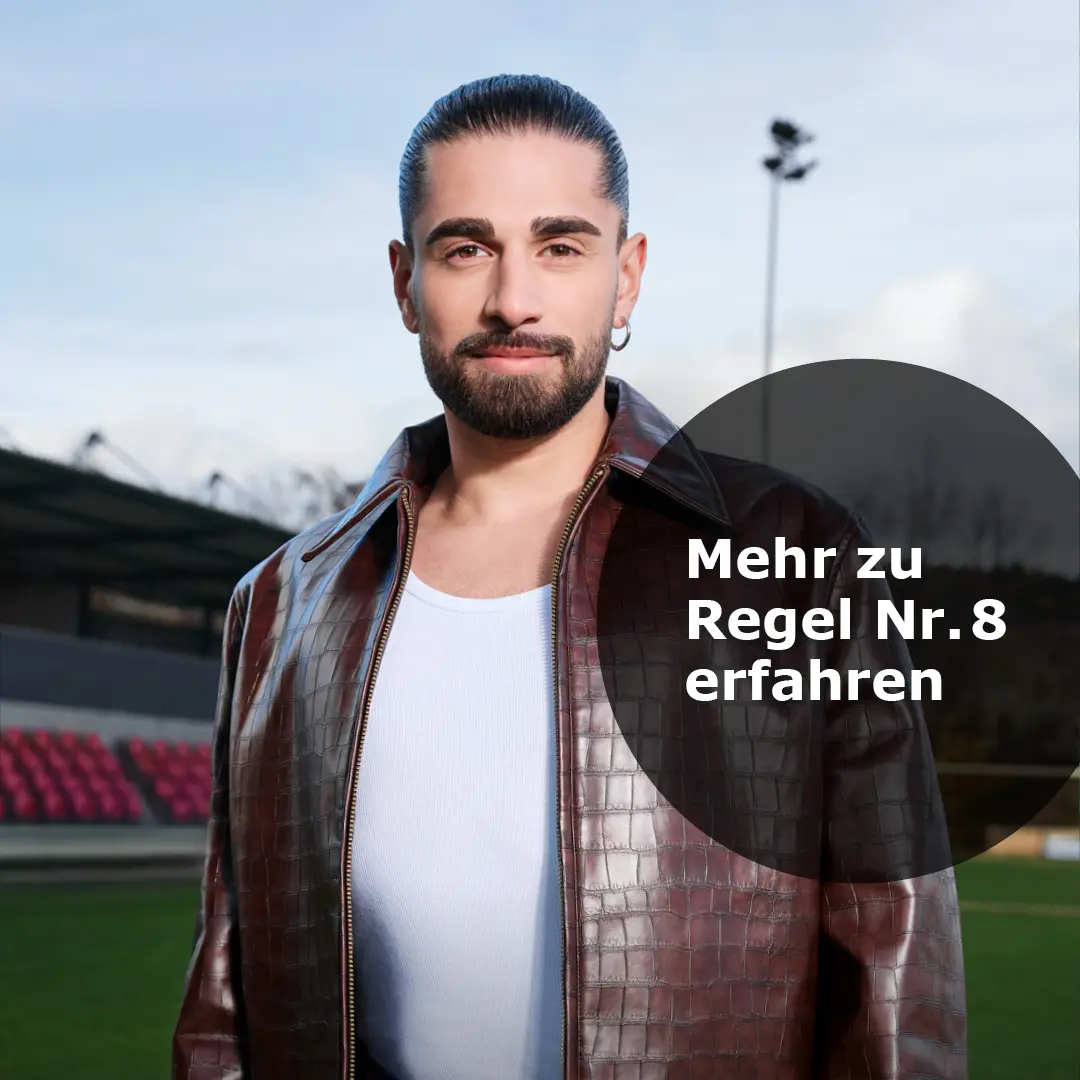 Porträt eines Mannes mit gepflegtem Bart und zurückgebundenem Haar, der eine braune Lederjacke und ein weißes Shirt trägt, im Hintergrund ein Stadion mit grüner Wiese und roten Sitzen.